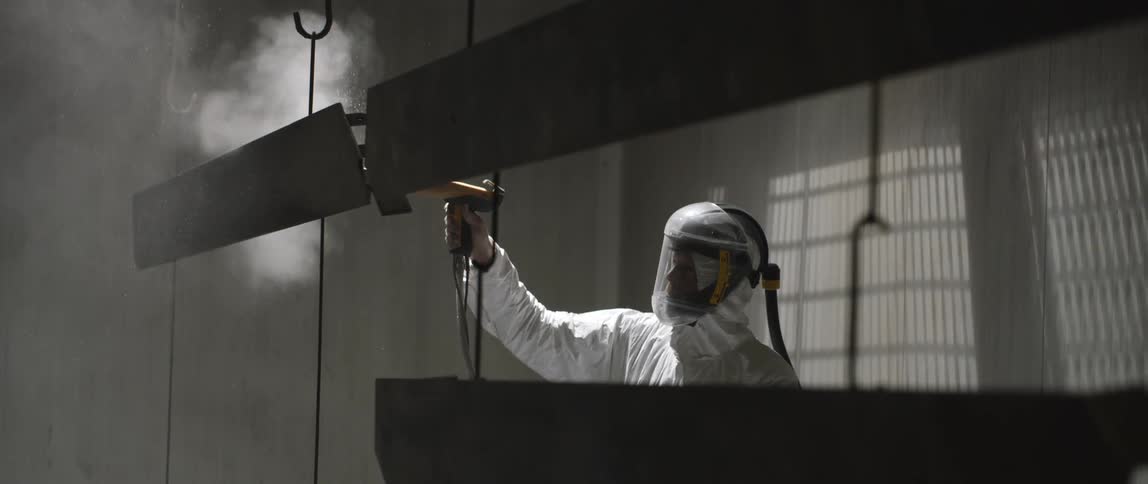 A person in protective gear using a tool to spray metal slabs suspended from the ceiling by hooks