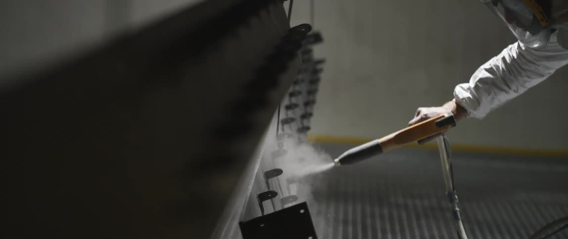 A man wearing a face shield and coveralls uses a power sprayer on a suspended piece of metal in a workshop