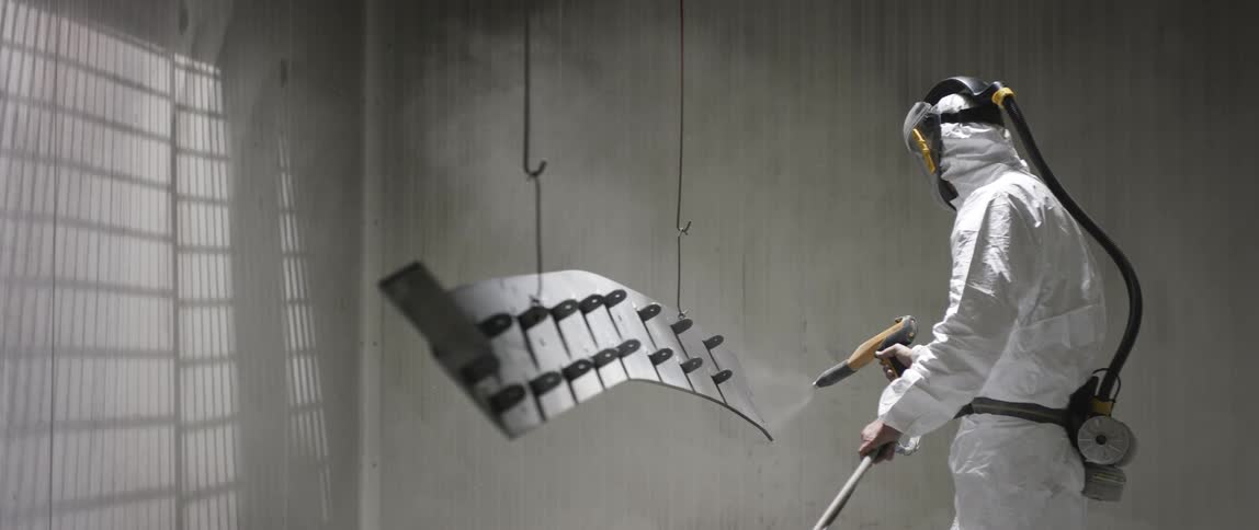 A person in protective gear using a power tool to spray a metal object hanging from a pair of hooks