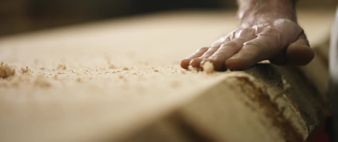 A white brushes shavings from a plank of wood with his hand