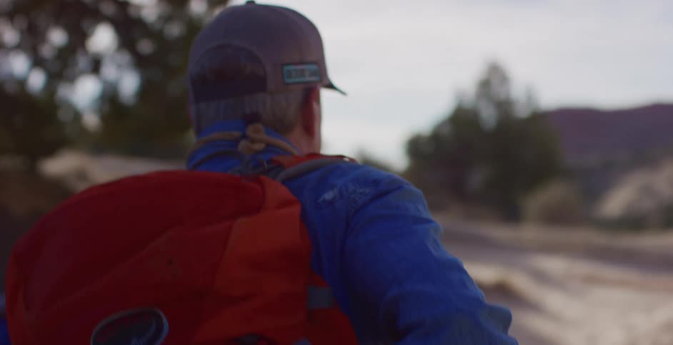 A man slings his orange backpack over his shoulder and puts it on as he hikes a trail