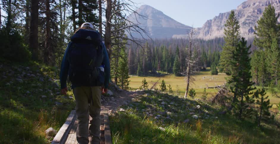 Male and female carry hiking backpacks as they walk along a brick pathway through a wooded area that leads to an open grassy plain beneath the tall mountain range on a bright sunny day