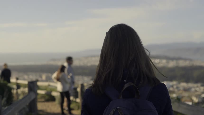 A woman with a backpack walks along a scenic route overlooking a city