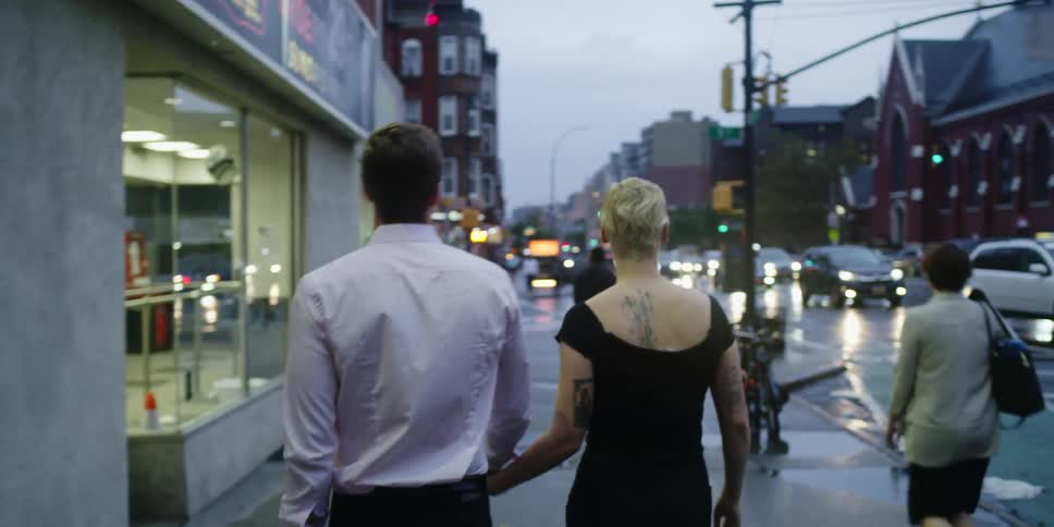 Male and female couple walk along the sidewalk in a small town holding hands at dusk as pedestrians and cars pass by