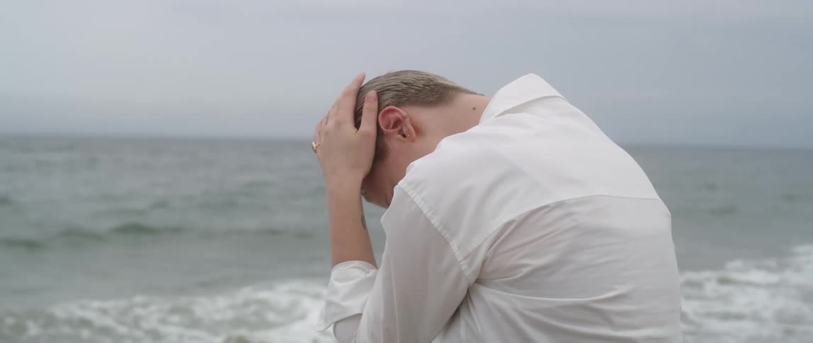 Woman leans head into hands in front of ocean and grey overcast sky