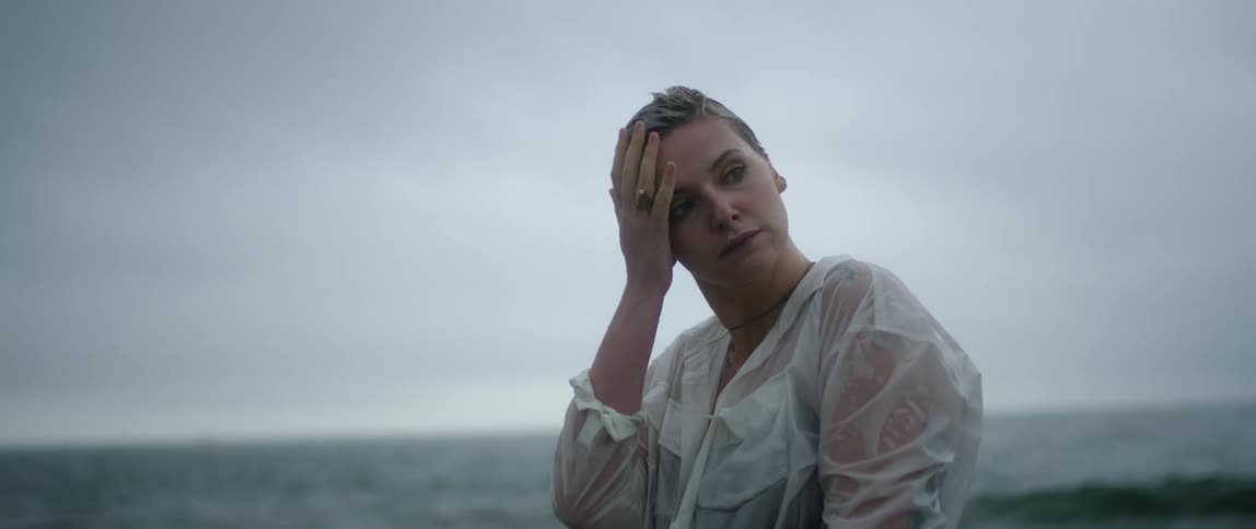A white woman in a raincoat sits on a beach