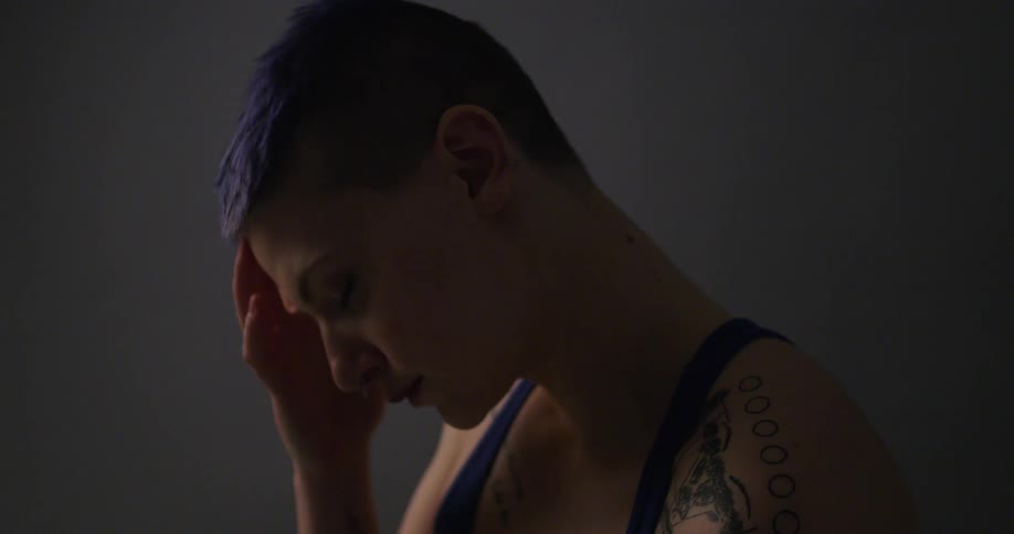 A woman with shorthair dyed blue stands in profile and lifts her hand to rub her head