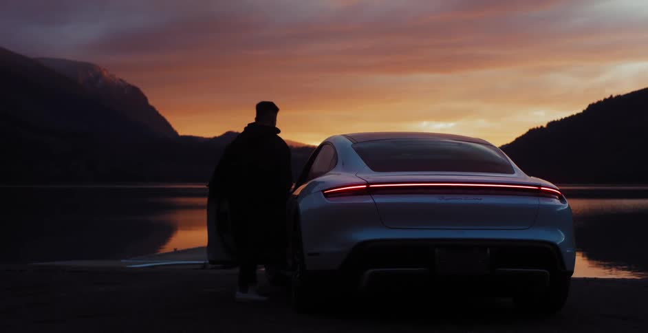 Man gets into car parked on shore with orange sky and mountains on the horizon