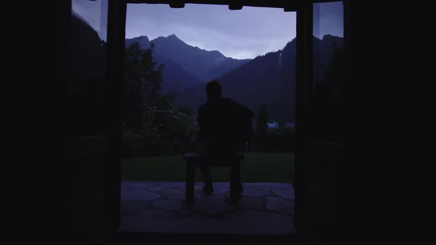 A man plays guitar and sings while sitting on a bench in view of the mountains