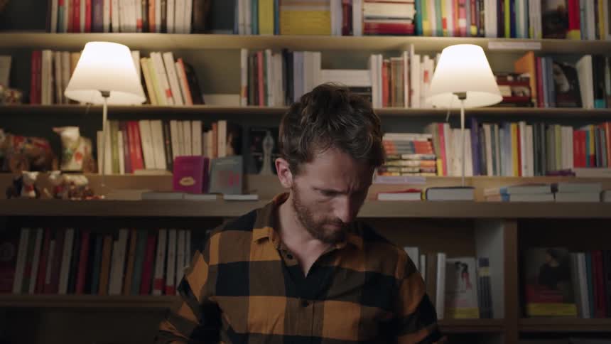 A man turns the pages of a book in a room with book shelves and lamps behind him