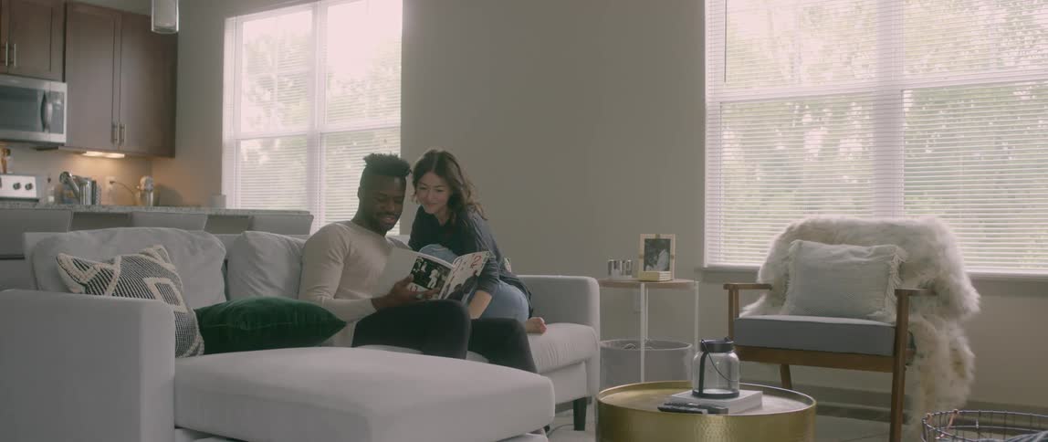 Male and female couple sit closely together on couch in living room as they talk about the magazine they are looking at together
