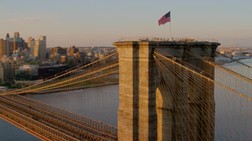 An american flag flies on top of a suspension bridge in a city