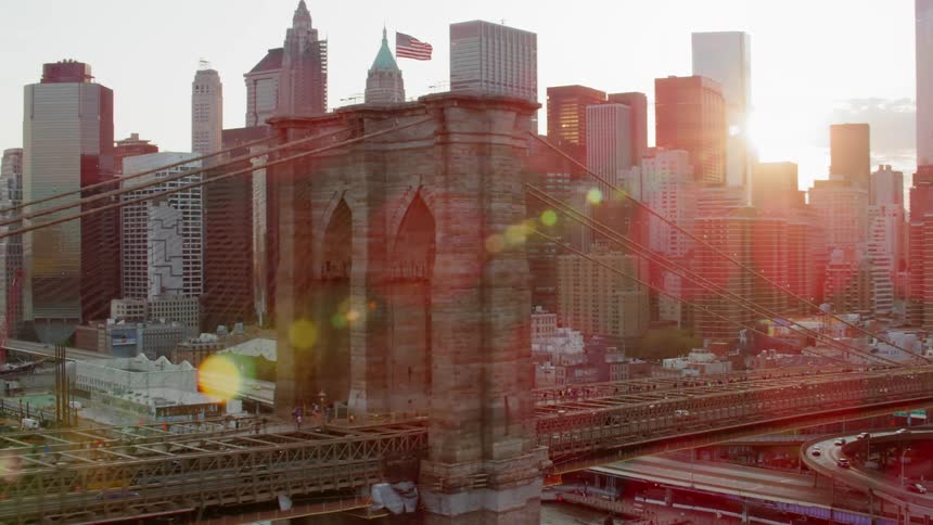 Cars drive over a large city bridge with skyscrapers in the distance at sunset
