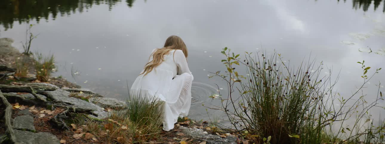 Woman with long hair on a white dress drinks water from a lake in the forest