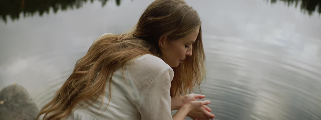 A woman drinks pond water from her cupped hands then turns and looks back over her shoulder
