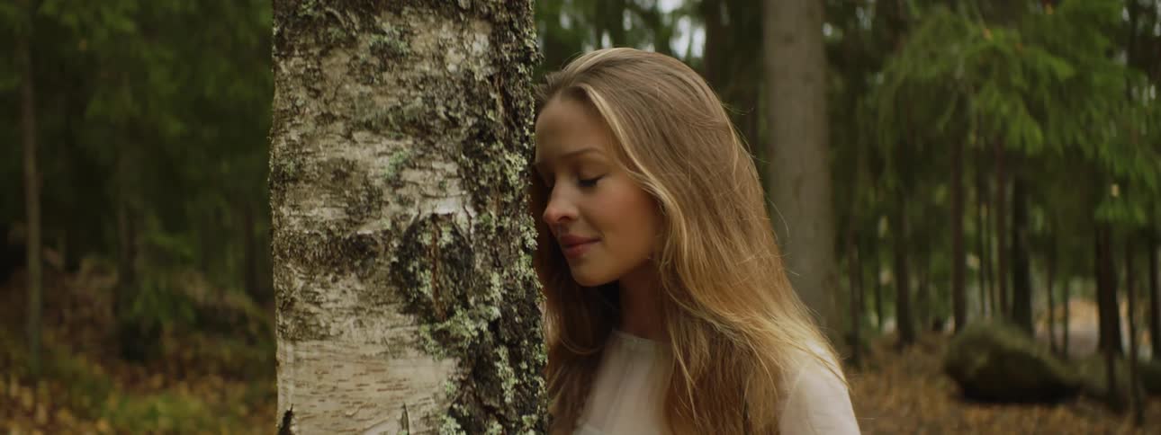 A white woman with long hair leans against a tree and then turns and walks away