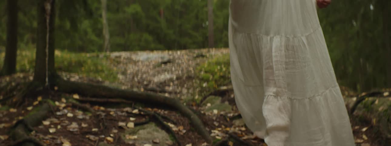 A woman wearing a white dress walks through a forest barefoot