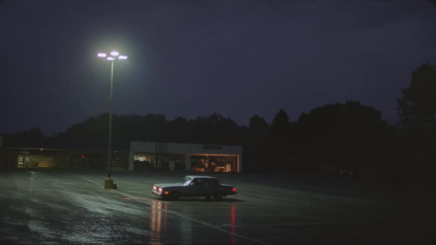 A car drivings and parks in an empty parking lot at night as it rains