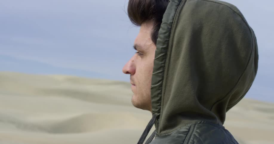 A man in a hoody standing among sand dunes in a desert blinks several times before closing his eyes