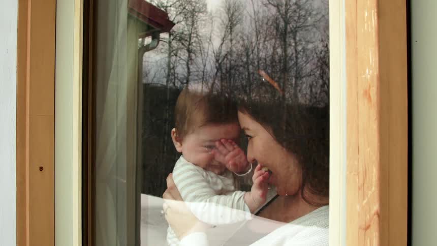 A woman looks out the window while holding her baby and laughing with him