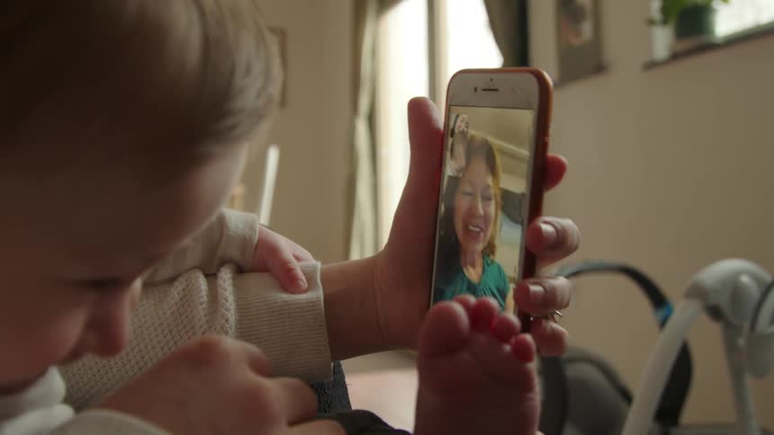 A mother holding her child in her lap while using a smartphone to video chat with an older woman