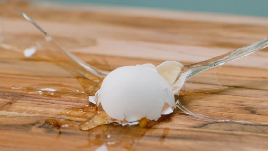 An egg falling onto a wooden cutting board and breaking causing egg yolk to splash out in every direction