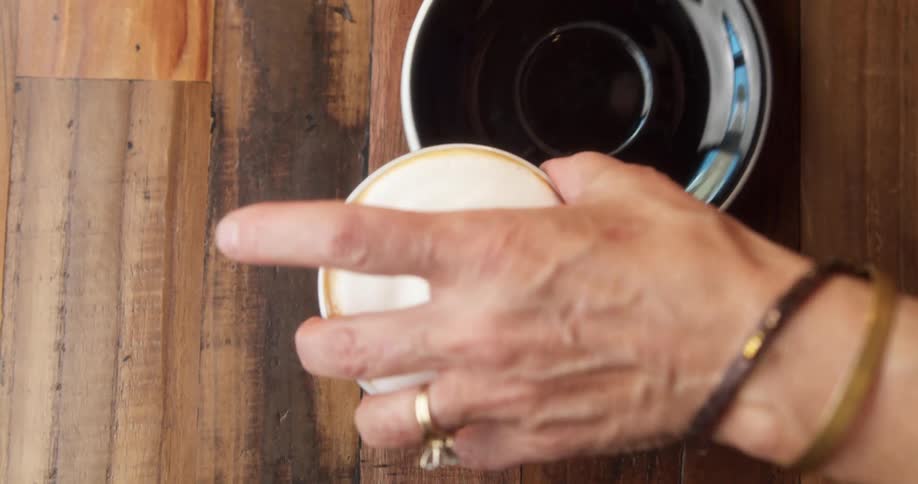 A white woman slides a saucer under a cup of coffee with a foamy head