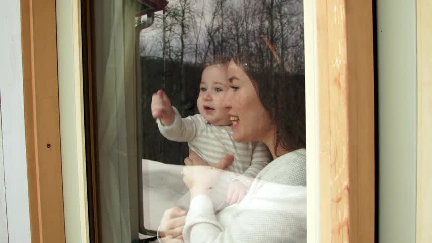 A woman holds a baby at a door with a window and the baby waves toward the outside view