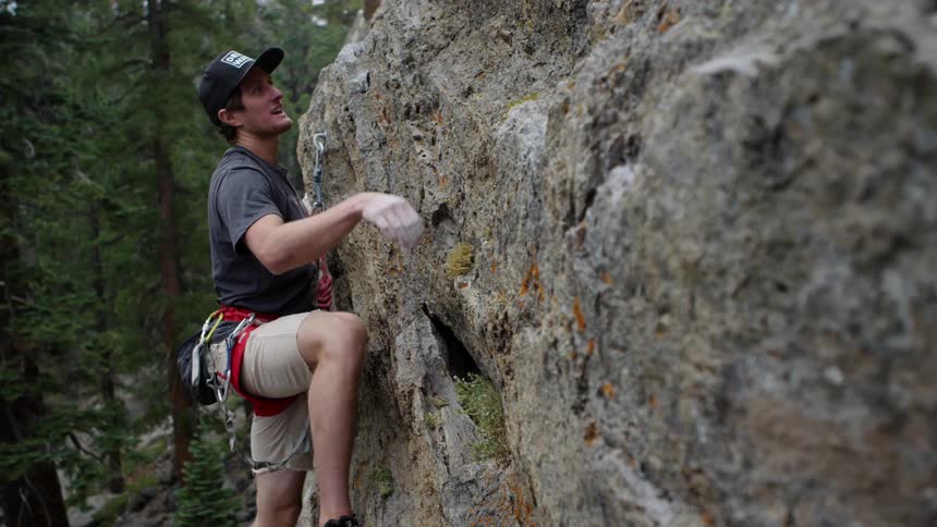 A man wearing a harness and a rope climbs up a rock face in the woods