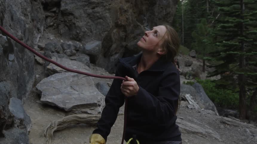 A white woman in climbing gear moves a rope while looking up the face of a mountain cliff