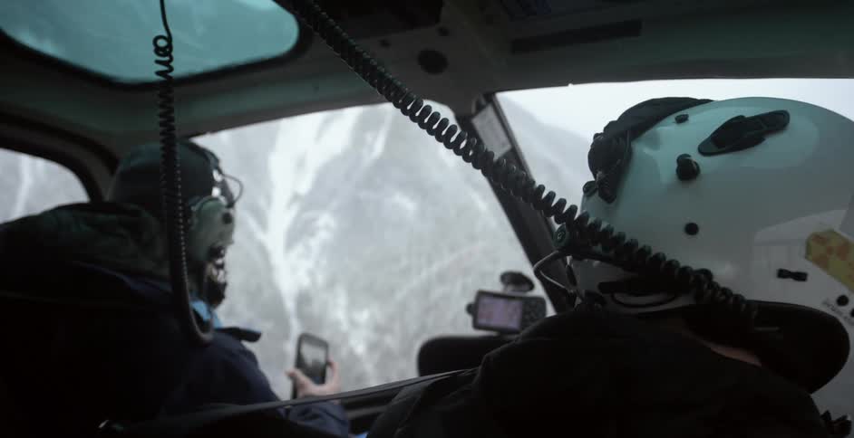 Two people in a helicopter flying over a snowy mountain