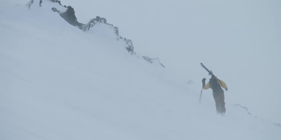 A skier walking on a windblown mountain holding skis