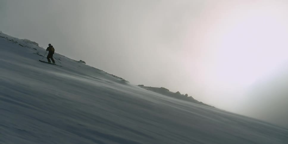 A skier moves across a windy mountain slope at sunset