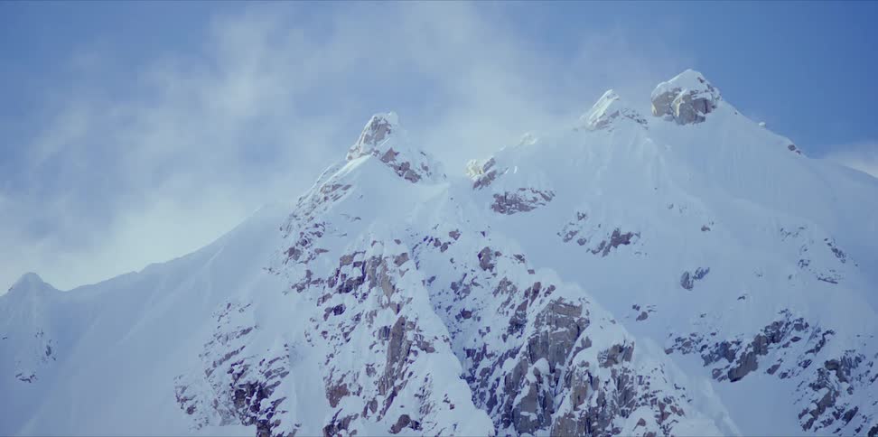A cloud moves over a snow covered mountain ridge on a bright blue sky day