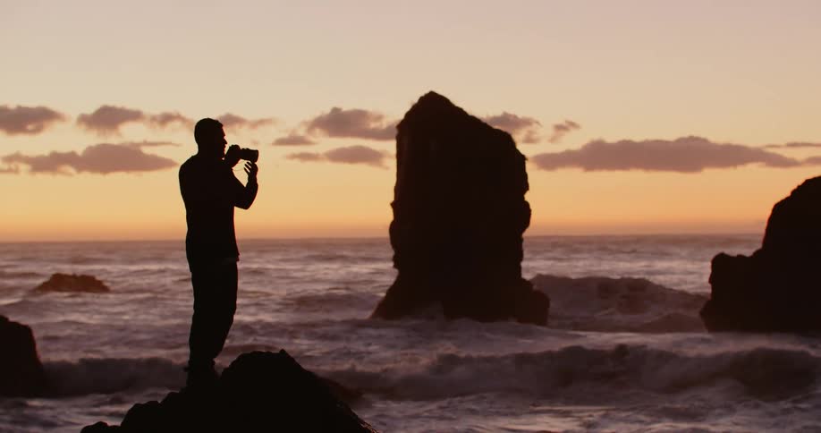 Man stands silhouetted on beach on rocks above surf taking photograph