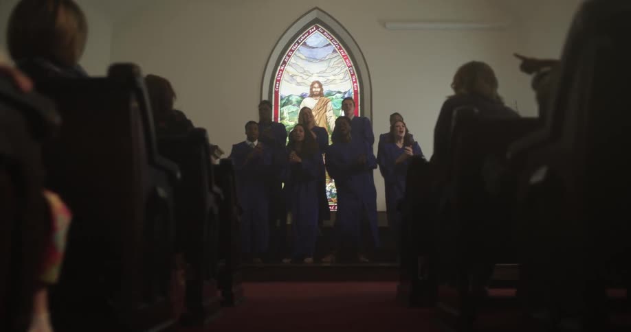 A choir singing and clapping with a stained glass window of jesus in the back