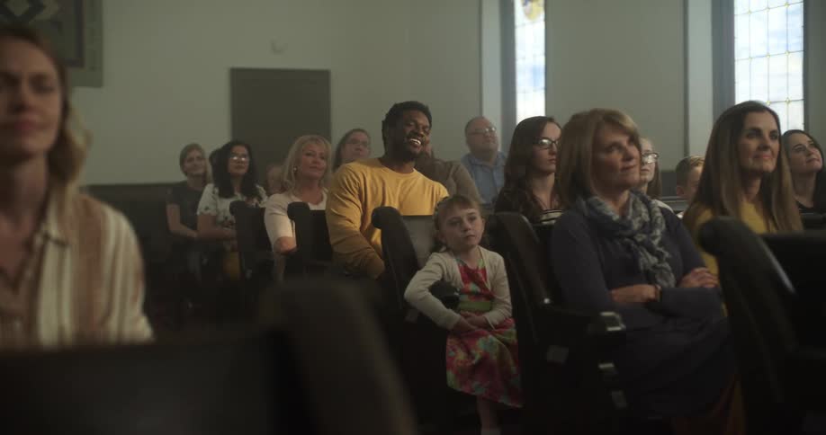 A black man laughs while sitting on pews in a church with other parishioners