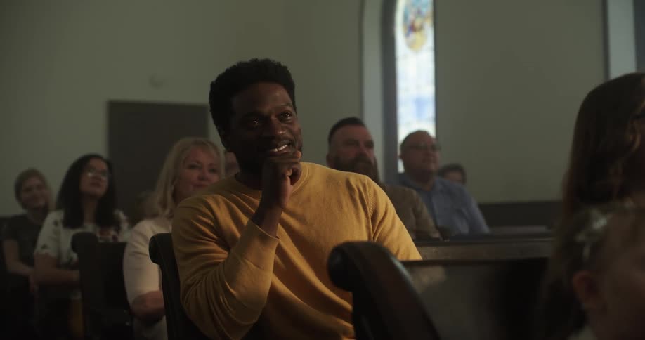 A black man sits in a pew at church with other parishioners and smiles