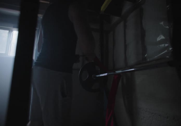 A man ties an athletic band around a bar weight in a dimly light garage gym