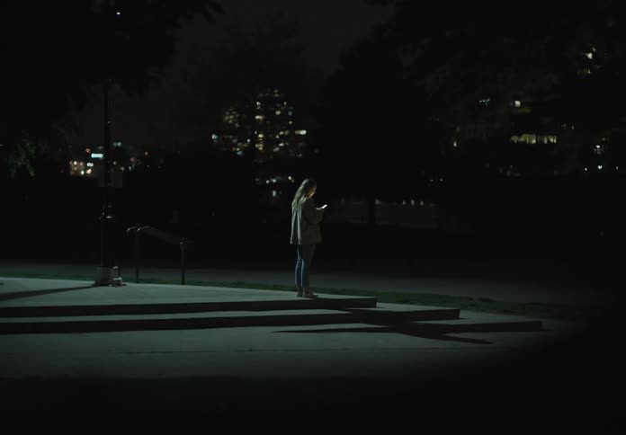 A woman stands on a step holding a phone and looking at it at night
