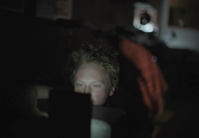 A white male sits in a dark room talking on a smartphone