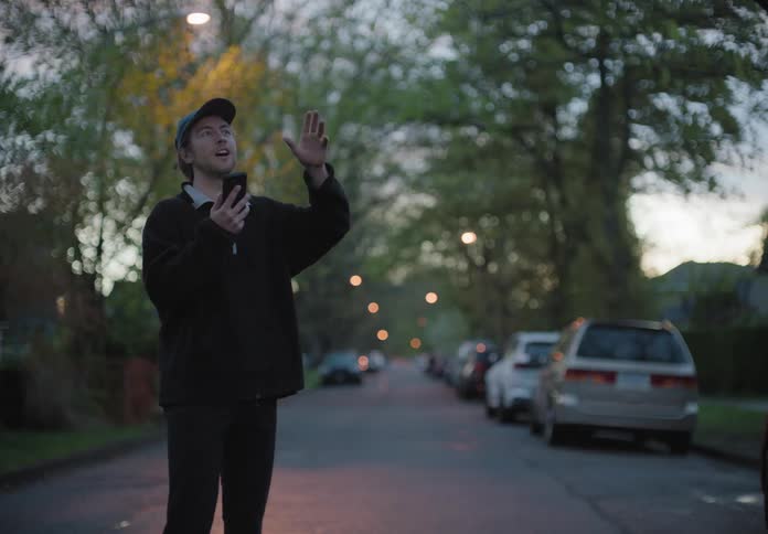A white man stands in the middle of a neighborhood street excitedly talking on a smartphone