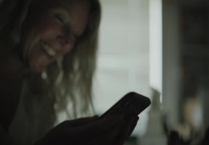 A woman sits in a house and smiles while using her smartphone