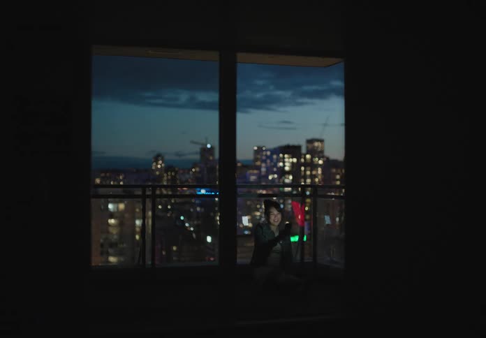 A young girl sits in front of a window with the city skyline behind her and video chats on her cell phone