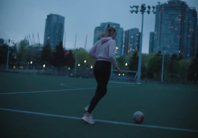 A white teenage kicks a soccer ball across an outdoor soccer field in the city