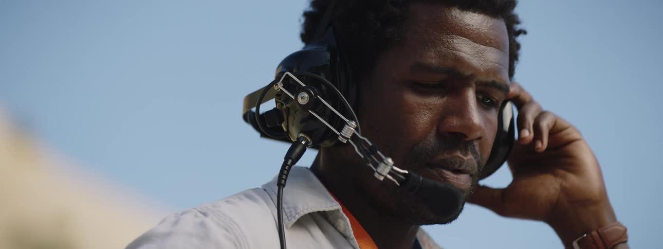 A black man stands outdoors talking into a headset