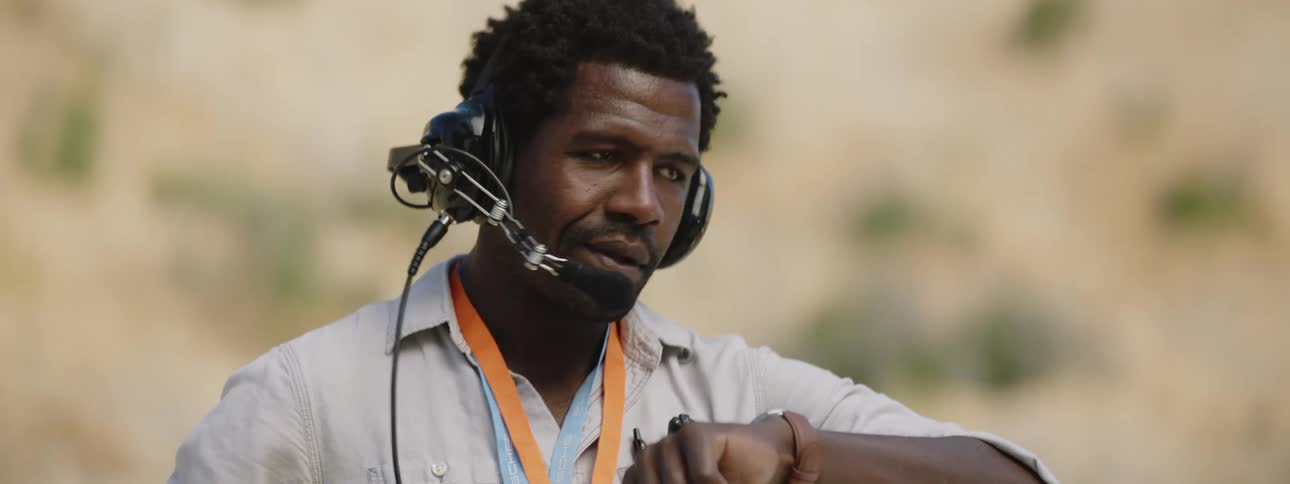 A man stands looking at his watch wearing a radio headset and shaking his head as he waits
