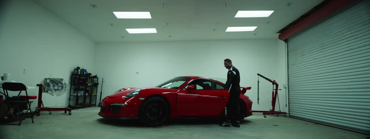 A man in a racing jumpsuit turns on the lights in a garage and gets into a red porsche