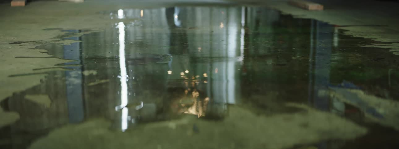 A man uses a tool to work on a vehicle on a lift in a garage while sparks fly towards a puddle on the floor