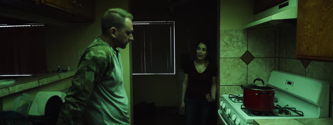 Female leans against the doorway to the kitchen as she has a serious discussion with male as he leans against the kitchen counter listening with remorse